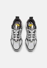 Schwarz-weiße Sneakers mit einem Mesh-Obermaterial, reflektierenden Akzenten, klobiger Form und einem auffälligen Schnürsystem. Gelbe Details auf den Innensohlen.