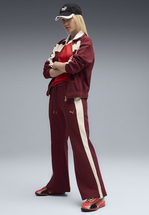 Chándal granate con detalles en crema y una gorra negra. Diseño deportivo con chaqueta con cremallera, pantalones de corte holgado y zapatillas rojas con detalles amarillos.