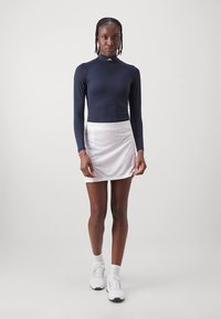 Tengerészkék hosszú ujjú felső magas nyakkal, finom mintákkal díszített fehér szoknyával párosítva. A modell fehér tornacipőt visel.