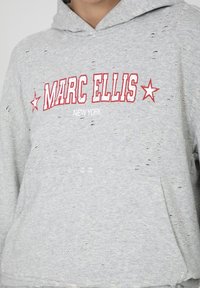 Felpa grigia in cotone decolorato con testo grafico rosso "MARC ELLIS" e "NEW YORK", dotata di tasca a marsupio e texture del tessuto visibile.