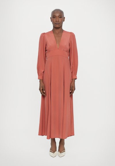 Langes, fließendes Kleid in Koralle mit tiefem V-Ausschnitt, Puffärmeln und tailliertem Schnitt. Weicher Stoff und knöchellanges Design. Helle, spitze Schuhe.