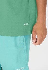 Grøn bomulds t-shirt med lige kant, der har et lille hvidt label. Sammen med lyseblå shorts, der viser en glat tekstur og syningsdetaljer.