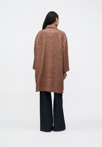 Pessoa com cabelo longo e cacheado vestindo um casaco solto de tom castanho texturizado e calças largas de tom azul escuro, de pé virada de costas contra um fundo simples.