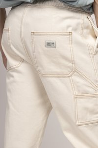 Pantalons en coton couleur crème avec des accents cousus et des poches visibles. Dispose d'une étiquette avec le logo "KAOTIKO SOCIETY" sur la poche arrière.