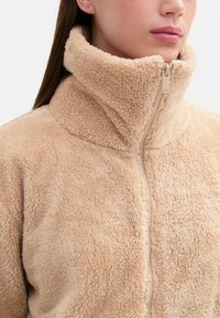Chaqueta de forro polar con cremallera en beige claro, con cuello alto, textura suave y una cremallera central. Diseño minimalista, sin patrones ni acentos adicionales.