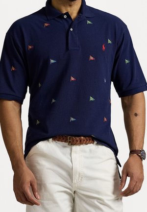 Marineblå polo shirt med små multifarvede flagmønstre, rød logo, kombineret med hvide bukser, brunt flettet bælte og ur på venstre arm.