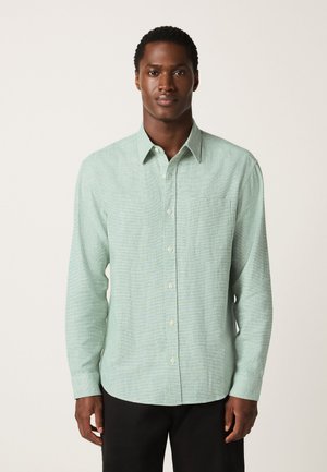 Homme portant une chemise boutonnée à manches longues vert clair et un pantalon noir, debout devant un fond clair uni.
