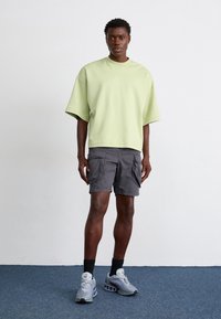 Ung man som står på en blå matta, bär en ljusgrön oversized kortärmad topp, mörka cargo-shorts, svarta strumpor och grå sneakers.