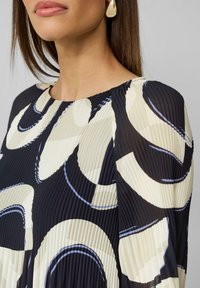 Haut plissé noir et crème avec un motif en vagues, doté d'un col rond et d'un tissu lisse et léger. Des boucles d'oreilles en forme de goutte d'or soulignent l'ensemble.