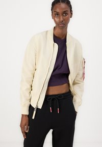 Veste bomber crème avec poignets côtelés, fermeture éclair et poche latérale. Portée sur un crop top violet et un pantalon de jogging noir avec cordon de serrage.