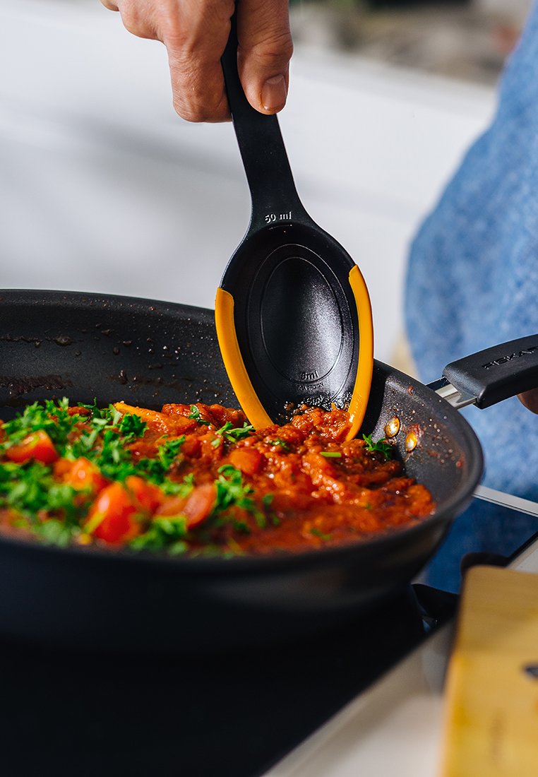 Schwarzer Spatel mit orangefarbener Akzent, der Sauce aus einer Pfanne mit gehackten Tomaten und Kräutern auf dem Herd schöpft.