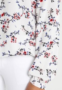 Blouse blanche à manches longues avec un motif floral en rouge, violet, rose et noir, associée à un pantalon blanc ; le poignet de la manche est élastiqué.