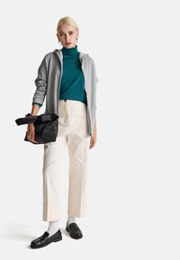 Graue Zip-Hoodie über einem türkisfarbenen Rollkragenpullover, kombiniert mit cremefarbenen Hosen und schwarzen Slippern. Hält eine schwarze Tasche mit strukturierten Details.