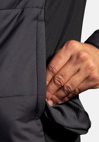 Giacca nera in tessuto morbido, con una tasca laterale visibile a cui si accede con una mano. Il design sottolinea una silhouette slanciata.