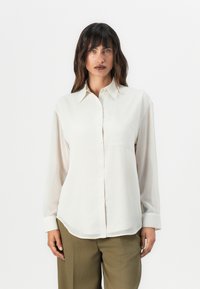 Light beige button-up shirt with a collared neck, long sleeves, front pocket, and slightly sheer fabric. Smooth texture and relaxed fit.