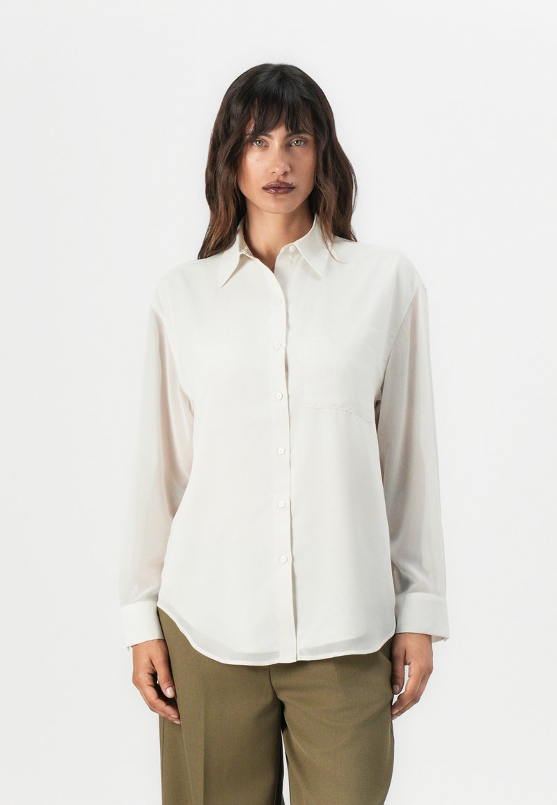 Chemise boutonnée beige clair avec un col, des manches longues, une poche avant et un tissu légèrement transparent. Texture lisse et coupe décontractée.