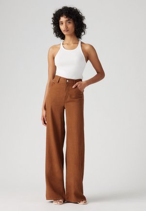 Calças largas castanhas feitas de tecido suave, com uma cintura alta, fecho de botão frontal e bolsos laterais, combinadas com uma t-shirt branca de alças.