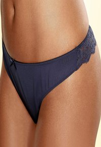 Marineblauwe string met kantafwerking, van gladde stof, met een lage taille en een delicaat strikje aan de voorkant. Heeft minimale stikdetails.