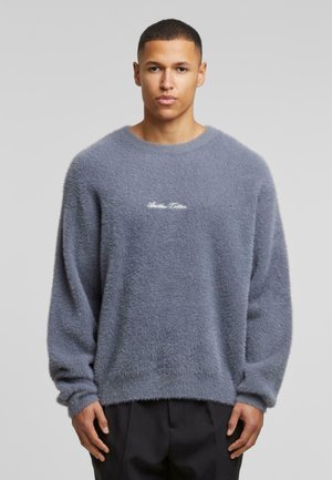 Suéter gris esponjoso de cuello redondo con mangas largas, que presenta una superficie texturizada y un logo bordado en blanco en el pecho.