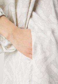 Garbamento leggermente testurizzato e decorato in morbido bianco e grigio chiaro. Presenta una tasca laterale con un sottile design geometrico.