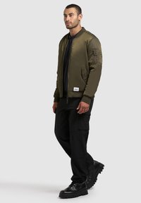 Olivgrüne Bomberjacke mit geripptem Kragen und Bündchen, Reißverschluss und strukturierten Schulterdetails, kombiniert mit schwarzen Cargo-Hosen und Stiefeln.