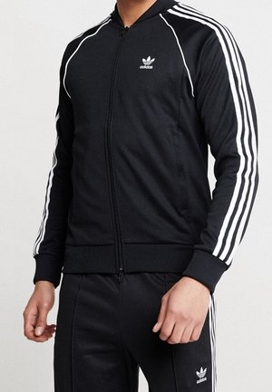 Osoba ubrana w czarny dres Adidas z białymi paskami na rękawach i nogawkach, stojąca na tle jasnego, jednolitego tła.