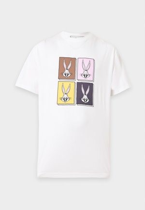 Mężczyzna w białej koszulce z czterema kolorowymi nadrukami Bugs Bunny, luźnych brązowych spodniach i czarnych trampkach z białą podeszwą stoi na jednolitym tle.