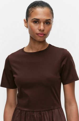 Kvinde med glat sammenbundet hår, iført små øreringe i ringform og en mørkebrun kortærmet top mod en ensfarvet hvid baggrund.