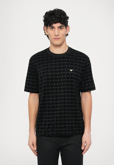 Schwarzes kurzärmliges T-Shirt mit einem strukturierten Muster aus ineinandergreifenden Dreiecken, rundem Ausschnitt und einem kleinen goldenen Emblem auf der Brust.