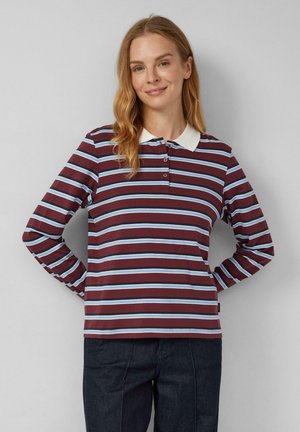 Vrouw met lang blond haar, gekleed in een gestreept polo-shirt met lange mouwen in bordeauxrood, blauw en wit, gecombineerd met donkere broek, staande met de handen achter zich.