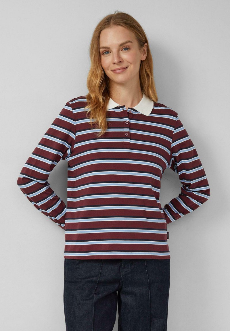 Vrouw met lang blond haar, gekleed in een gestreept polo-shirt met lange mouwen in bordeauxrood, blauw en wit, gecombineerd met donkere broek, staande met de handen achter zich.