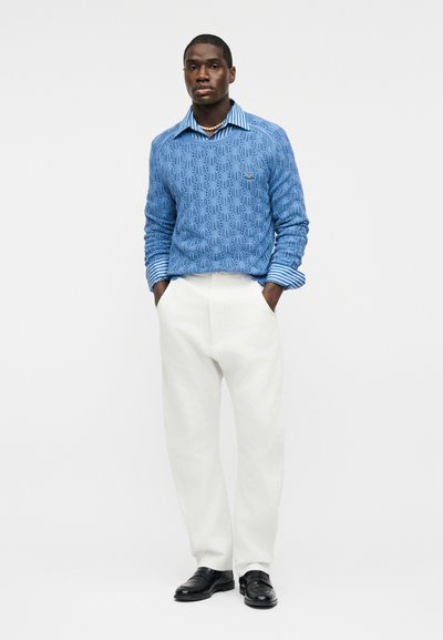 Homme portant un pull texturé bleu sur une chemise à rayures bleues, un pantalon blanc, des chaussures noires et un collier de perles, debout les mains dans les poches.