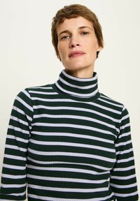 Frau mit kurzen braunen Haaren, die einen dunkelgrünen und weißen gestreiften Rollkragenpullover trägt und frontal vor einem schlichten Hintergrund steht.