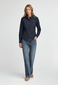 Camicia button-up blu scuro con maniche lunghe, il tessuto è liscio. Abbinata a jeans a vita alta sbiaditi e scarpe slip-on marrone chiaro.