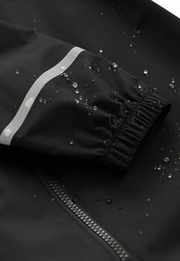 Chaqueta impermeable negra con una franja reflectante plateada, puños elásticos y gotas de agua en la superficie, destacando su resistencia al agua.