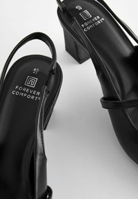 Sandalias de tacón negro con un acabado brillante, que cuentan con una correa trasera y un tacón ancho. Logotipo "Forever Comfort" visible en la plantilla.