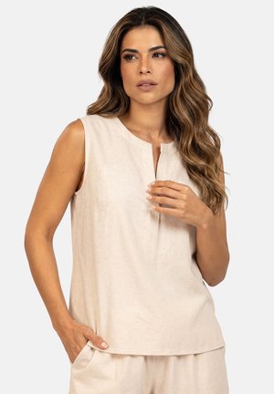 Kobieta o długich falowanych włosach, ubrana w beżowy bezrękawnik i pasujące spodnie, jedna ręka w kieszeni, druga przy piersi, neutralne tło.