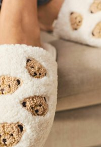 White plush slippers featuring embroidered bear faces in beige and brown. Soft texture with an open-toe design, ideal for indoor wear.