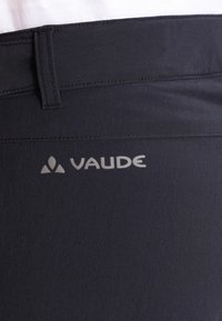 Černé látkové kalhoty s hladkým povrchem, zašitou elastickou gumou v pase a reflexním logem VAUDE na levé straně.