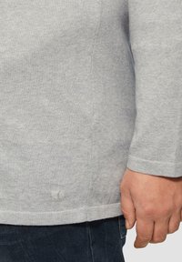 Grauer Sweatshirt aus strukturiertem Stoff, mit gerippten Bündchen und Saum. Verfügt über ein kleines Logo nahe dem Saum. Hand hält den Stoff.