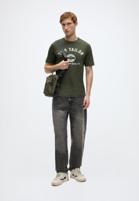 Πράσινο ελιάς T-shirt με λευκό κείμενο, γκρι ξεθωριασμένο τζιν, και κρεμ αθλητικά παπούτσια. Μικρή πράσινη τσάντα ελιάς με ιμάντα ώμου, με τσέπη με φερμουάρ.