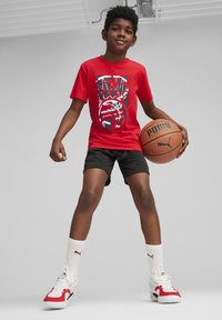 Camiseta roja con diseño gráfico, pantalones cortos negros, calcetines blancos y zapatillas rojas y blancas, sosteniendo un balón de baloncesto marrón, sobre un fondo gris.