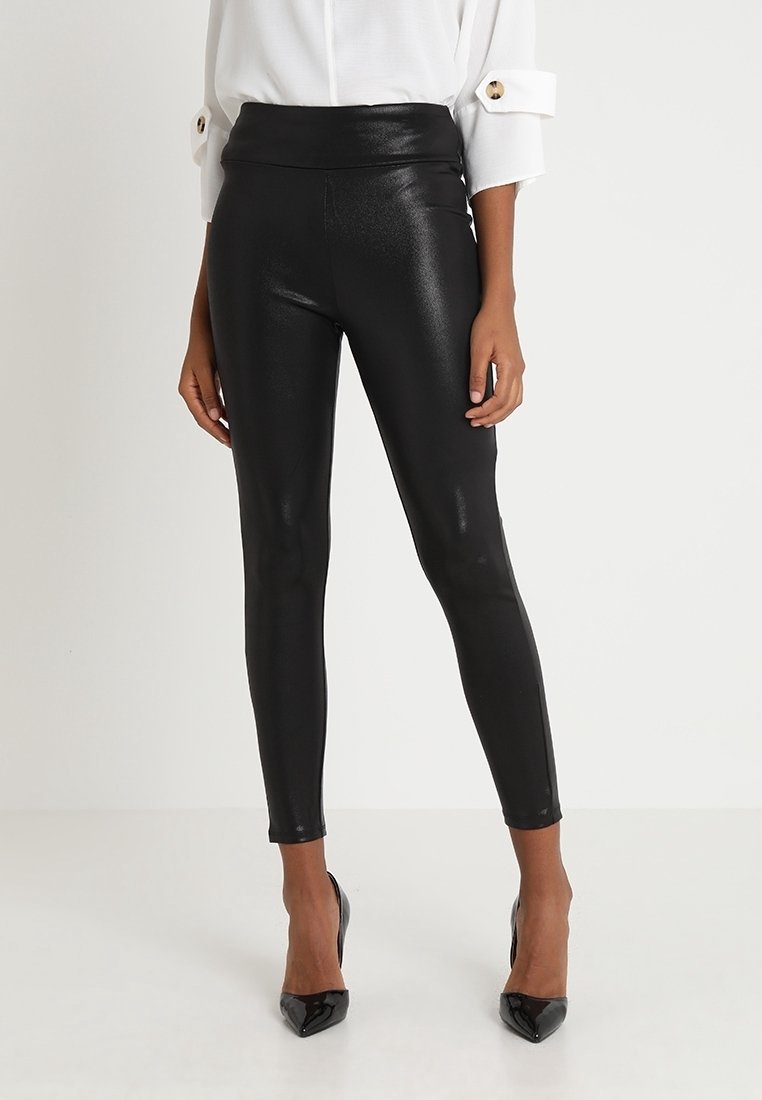 Femme portant des leggings noirs brillants et moulants, un chemisier blanc à manches longues avec poignets boutonnés, et des chaussures à talons noirs sur fond blanc.