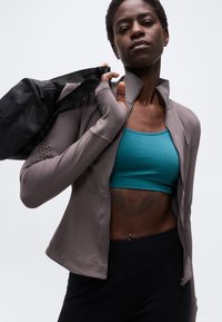 Αθλητική γυναίκα που φοράει πετρόλ αθλητικό σουτιέν και γκρι μπουφάν, κρατώντας μαύρη τσάντα γυμναστηρίου στον ώμο, με ορατό τατουάζ λιονταριού και σκουλαρίκι στον ομφαλό.