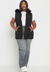 Mustat puffervestit, joissa on tekoturkkihuppu, vetoketjukiinnitys, sivutaskut ja tikattu muotoilu, käytetään valkoisen paidan ja harmaiden farkkujen päällä.