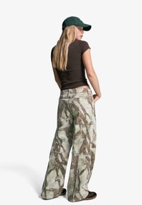 Smeđi uski top, kamuflažne cargo hlače s svijetlozelenim i smeškim uzorcima te zelena kapa. Opušteni kroj s bočnim džepovima i vidljivim šavovima.
