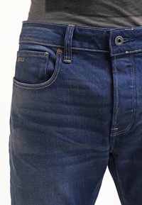 Mörkblå denimjeans med rak passform, femficksdesign och bronsdetaljer med synliga sömnadsdetaljer.