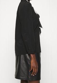 Schwarze Bluse mit langen Ärmeln aus weichem Stoff. Besitzt eine Schleife am Kragen und kontrastierende weiße Akzente. Kombiniert mit einem schwarzen Lederrock.