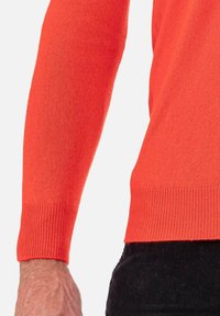 Pull en tricot orange avec un poignet côtelé et une texture lisse, montré partiellement sur un bras.