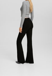 Mujer vistiendo un suéter gris de canalé ajustado y unos pantalones vaqueros negros de talle alto y acampanados, de pie contra un fondo sencillo.